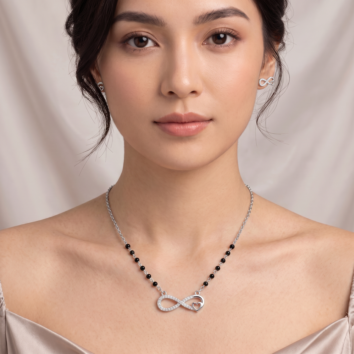 A woman wearing a delicate silver necklace with a silver infinity symbol pendant and small black beads (mangalsutra), paired with matching silver stud earrings