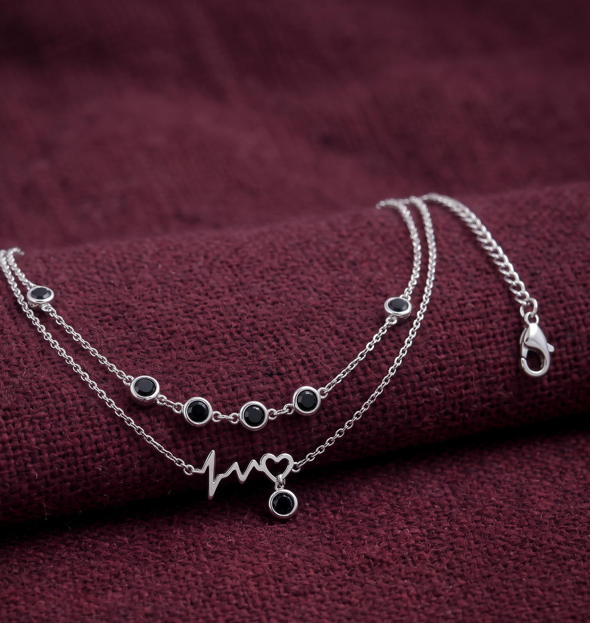 A silver double-layered Anklet with small round dark stones and a heartbeat line with a heart pendant