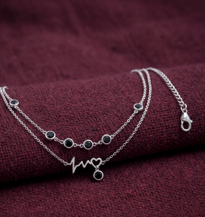 A silver double-layered Anklet with small round dark stones and a heartbeat line with a heart pendant
