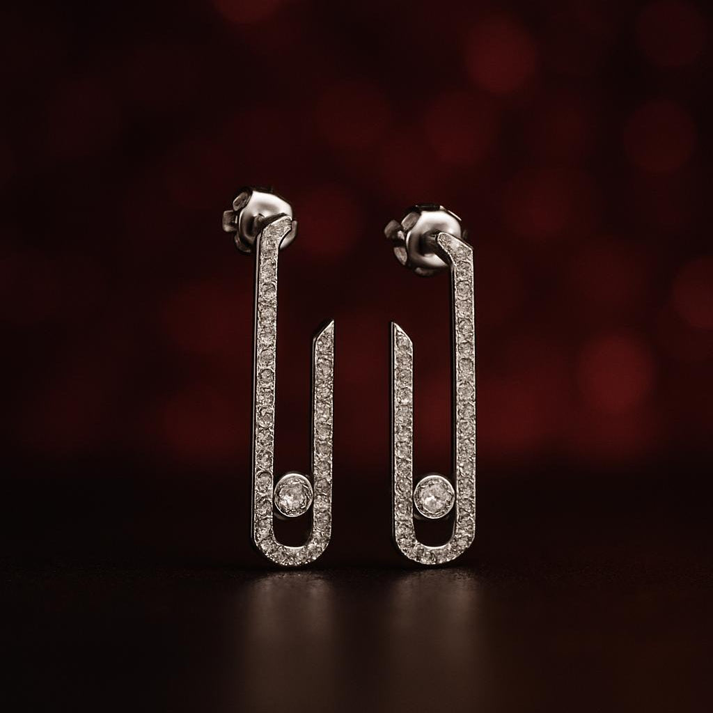 A pair of silver-toned safety-pin style earrings paved with small clear stones, standing upright on a dark reflective surface against a dark red bokeh background.