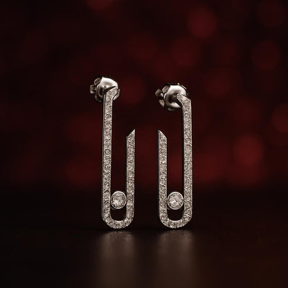 A pair of silver-toned safety-pin style earrings paved with small clear stones, standing upright on a dark reflective surface against a dark red bokeh background.