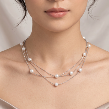 A close-up of a woman wearing a multi-layered silver necklace with small white pearls, set against a soft beige draped fabric background.